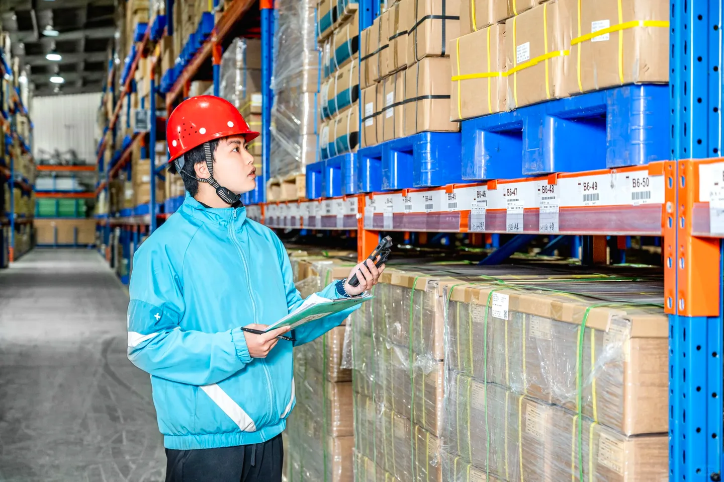 Warehouse worker checking boxed stock and records beside pallet racks